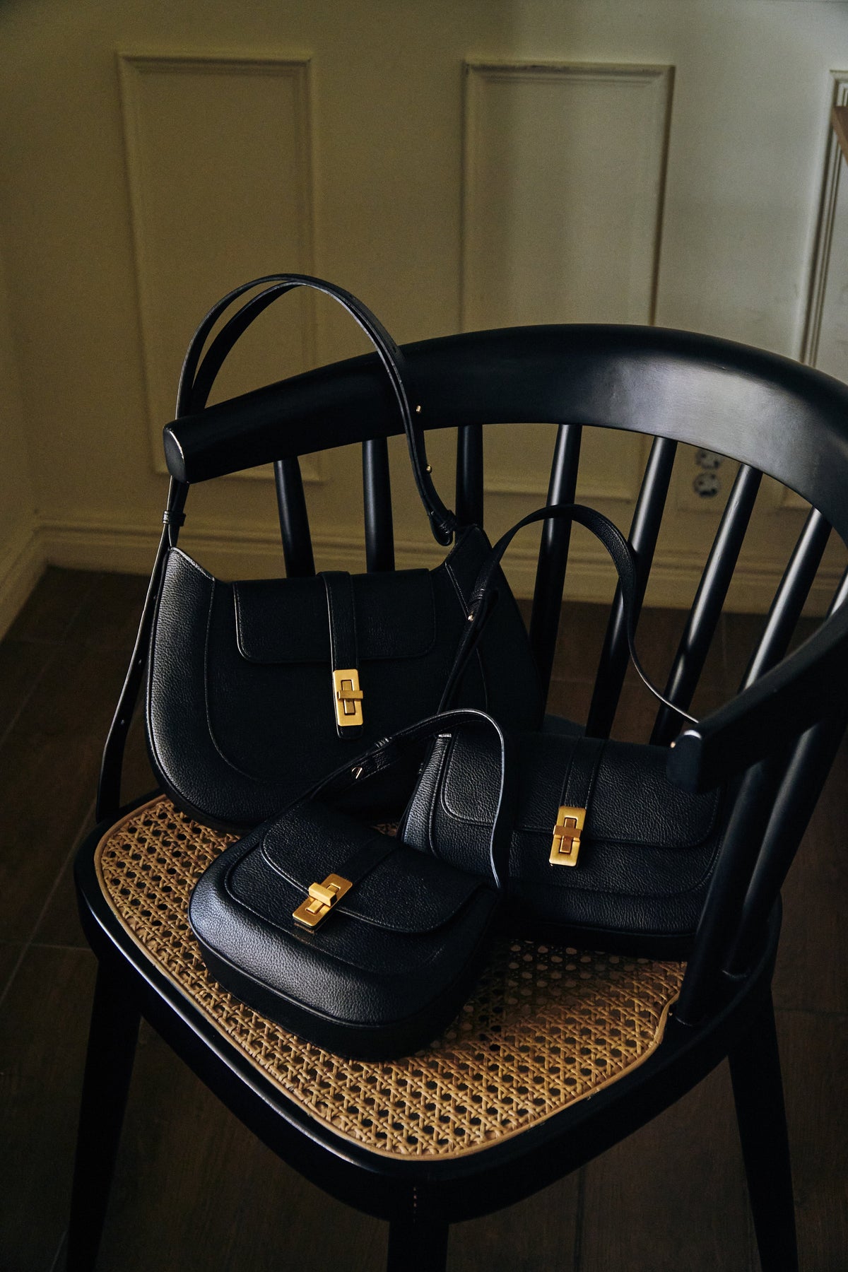 Three black MJade bags with gold hardware arranged on a cane chair.